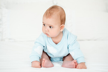 Fototapeta premium A baby boy in a blue bodysuit on a white bed at home learns to sit, a place and space for text, a healthy little child