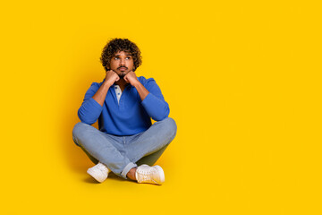 Young man sitting against a yellow background wearing a blue polo shirt and jeans, depicting casual fashion and thoughtful leisure