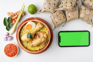 Blank green screen smartphone and Lontong opor ayam served during eid Mubarak, Eid al Fitr holiday celebration with Ketupat Lebaran or rice cake dumpling and sambal chili sauce.