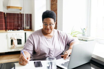 Portrait of elderly black plus size female studying at home sitting at kitchen table in front of laptop next to window, watching webinar, noting down tips and information after speaker
