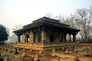 Obraz premium Pavilion at Shalimar Bagh with stone structures. Mughal architecture, cultural heritage, landscape photography. Formal garden, misty scene, Srinagar landmark, capturing the history of Mughal garden