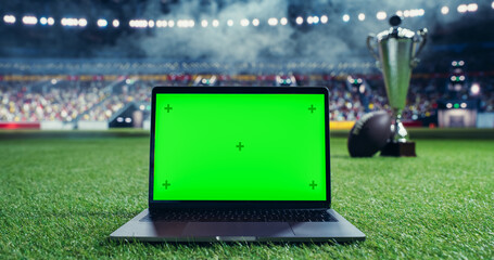 Laptop Computer with a Green Screen Mock Up Display Standing Next to an American Football and a Championship Trophy. Template for Software, Streaming, Sports Betting, Multimedia Content