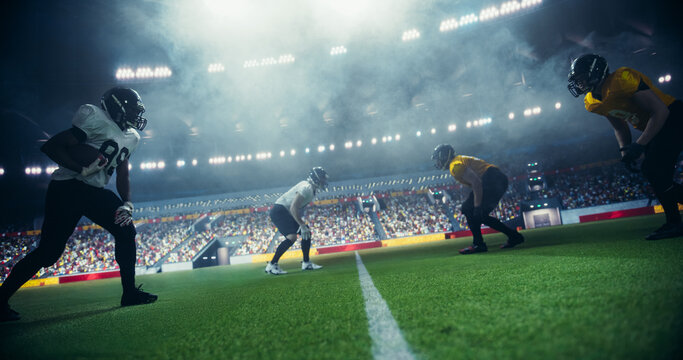 American Football Players Compete in a Stadium, Illuminated by Bright Lights With a Cheering Crowd in the Background. Athletes Play an Intensive Dynamic Game, Tacle, Try to Score a Touchdown