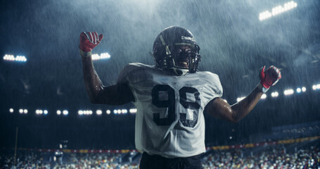 Portrait of Male Football Player Celebrating Scoring a Successful Touchdown Under Heavy Rain. Athletic American Footballer Jumping on a Stadium Field. Crowd Cheering In The Background © Gorodenkoff