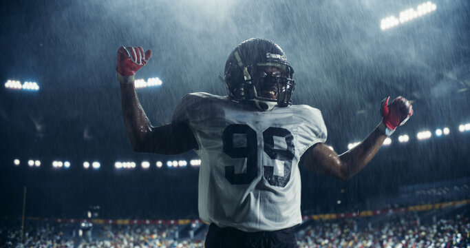 Portrait of Male Football Player Celebrating Scoring a Successful Touchdown Under Heavy Rain. Athletic American Footballer Jumping on a Stadium Field. Crowd Cheering In The Background