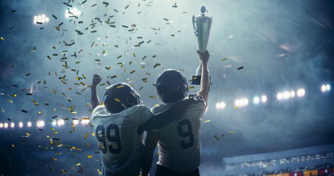 American Football Team Celebrating a Championship Victory with Golden Trophy and Falling Confetti Under Stadium Lights. Emotional Moment of Triumph for Two Diverse Footballers in White Jerseys - Powered by Adobe