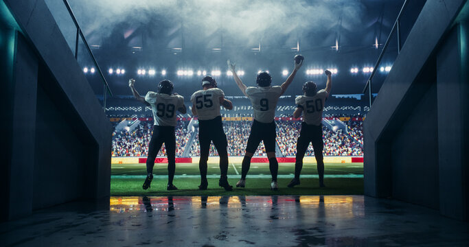 Diverse American Football Players Walking Out of a Stadium Tunnel, Raising Hands with Helmets, Preparing for Championship Game. Arena Crowd Cheering in the Background Under Bright Stadium Lights