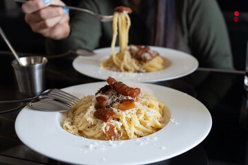 A plate of closeup carbonara spaghetti topped with grilled bacon with hand holding fork eating in background. It is an Italian pasta dish from Rome made with egg, hard grated cheese, cured pork.
