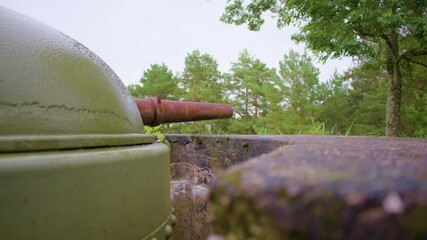 Panning shot of a weathered green turret with a rusted barrel emerging from a fortified concrete bunker, partially covered by overgrown grass and moss, blending into the surrounding dense forest.