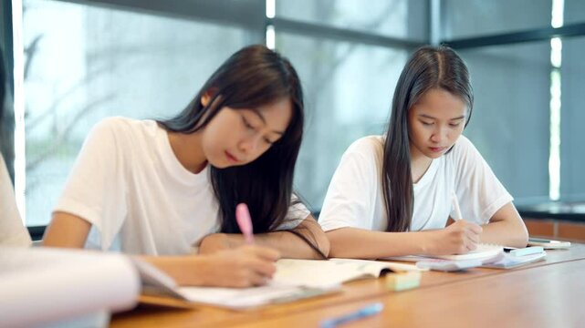 Two women are sitting at a table writing. One of them is wearing a white shirt. Scene is focused and studious