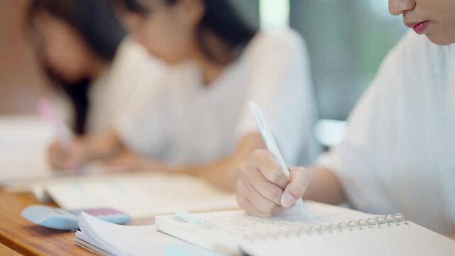 A girl is writing in a notebook with a pen. She is sitting at a desk with other girls. The girls are writing and seem to be focused on their work. Scene is serious and studious