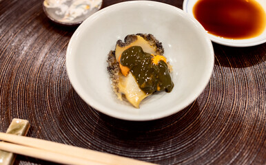 Top-down view of abalone (Awabe) sashimi topped with sea urchin, elegantly plated in a ceramic bowl with soy sauce on the side, showcasing the refined composition of Japanese cuisine