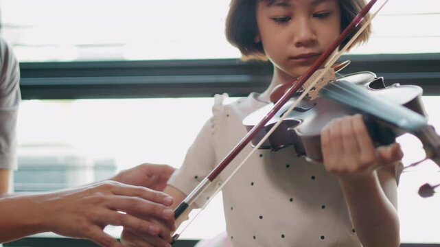 Asian young girl practices violin with focused dedication, guided by a supportive teacher showing music education, classical music, and the bond between student and mentor for nurturing talent.
