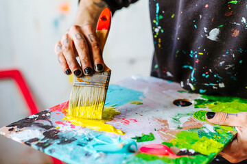 Artist's Hand Mixing Paint on a Color Palette. Close-up of an Argentine artist’s hand holding a brush covered in yellow paint, mixing colors on a palette during the creative process.