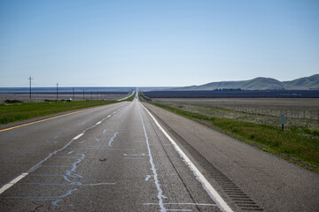 Das Bild zeigt eine weitläufige Wüstenautobahn, die sich unter strahlend blauem Himmel in die Ferne erstreckt. Auf der anderen Seite ein Feld aus trockenem Gras und Sträuchern. 