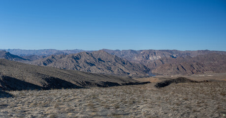 temberaubende Weite einer W&uuml;stenlandschaft unter strahlend blauem Himmel ein. Die Perspektive geht nahtlos vom lebendigen Vordergrund in das kahle, zerkl&uuml;ftete Gel&auml;nde &uuml;ber.