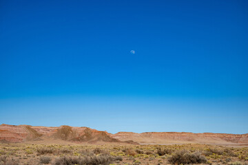 Das Bild zeigt eine weite, trockene Landschaft, die sich unter strahlend blauem Himmel. Ein Vollmond h&auml;ngt in der Ferne &uuml;ber einem offenen Feld, das zu einer Bergkette f&uuml;hrt. 
