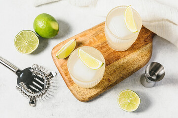 Refreshing summer lime cocktails on wooden board. Two glasses of citrus hard seltzer with ice cube, lime slices, surrounded by cocktail strainer and jigger. Lemonade, mocktail, caipirinha, spritz.