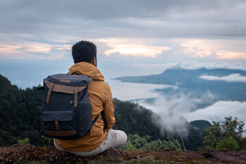 A traveler sits on a mountain peak, gazing at the misty valley and ocean at sunrise. Wearing a backpack and jacket, he enjoys the peaceful scenery, symbolizing adventure, freedom, and exploration.