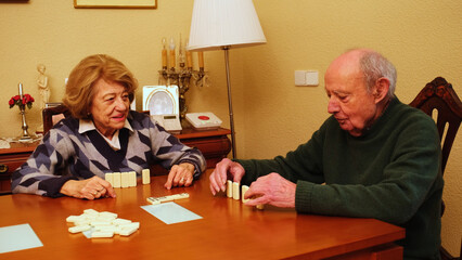Elderly couple playing dominoes on a wooden table in their living room, enjoying their free time...