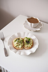 Aesthetic avocado toast on a white plate with a cup of coffee in a cozy, inviting atmosphere.