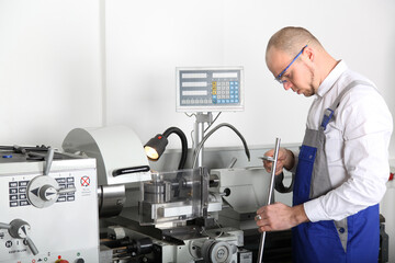 Lathe machine operator at work. A man in work clothes and safety glasses operates a lathe machine with a digital display in a workshop.