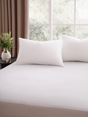 A bed with two pillows in a modern bedroom in front of a large window decorated with brown curtains. Bed is covered by a snow-white sheet. Close-up photo. Suitable for mockup