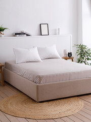 Modern bedroom with a minimalist interior, white walls, wooden floors and handcrafted furniture. Double bed with two pillows neatly covered with a fitted sheet. Side view photo. Suitable for mockup