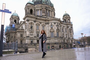 Fototapeta premium A stylish woman in a long coat walks along a pathway near Berlin Cathedral on a cold day, with scaffolding visible on the building.