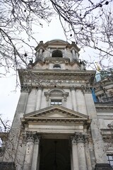 A dramatic low-angle view of Berlin Cathedral tower, framed by leafless branches, highlighting its intricate baroque architecture against a cloudy winter sky