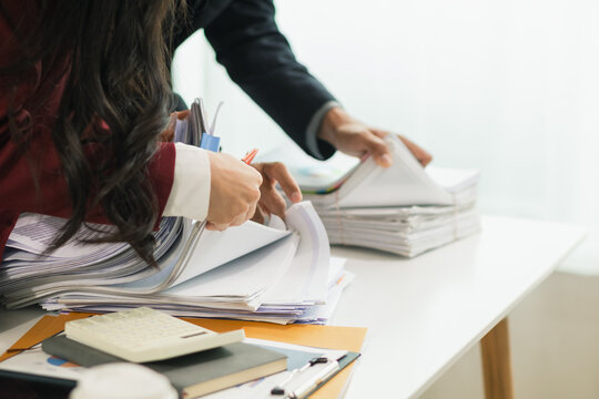 Two businesswomen are working together to search through important documents from a pile of documents on a table to verify the authenticity of the documents that were previously signed in a contract.