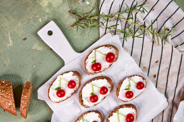 Creative tartines with white cheese and tomato cherries rest on a white board on a green table and a striped napkin