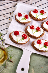 Creative tartines with white cheese and tomato cherries rest on a white board on a green table and a striped napkin