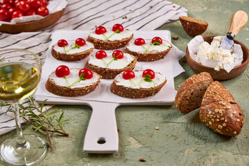 Creative tartines with white cheese and tomato cherries rest on a white board on a green table and a striped napkin