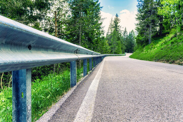 The image shows a lush green road curving through a forest near Danta di Cadore in Veneto, Italy, under a partly cloudy sky in June 2024