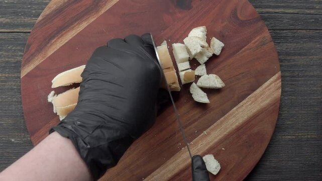 A knife slices a bun into small pieces, preparing them for baking crispy rusks in the oven. Close-up shot of precise cutting on a wooden board