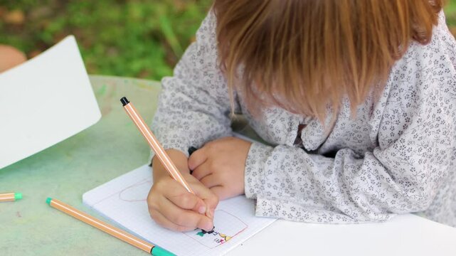A young child sits at a table in a summer garden, concentrating while drawing with a fineliner in a graph-paper notebook, expressing creativity through detailed sketch