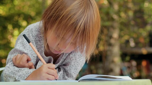 A young child sits at a table in a summer garden, concentrating while drawing with a fineliner in a graph-paper notebook, expressing creativity through detailed sketch
