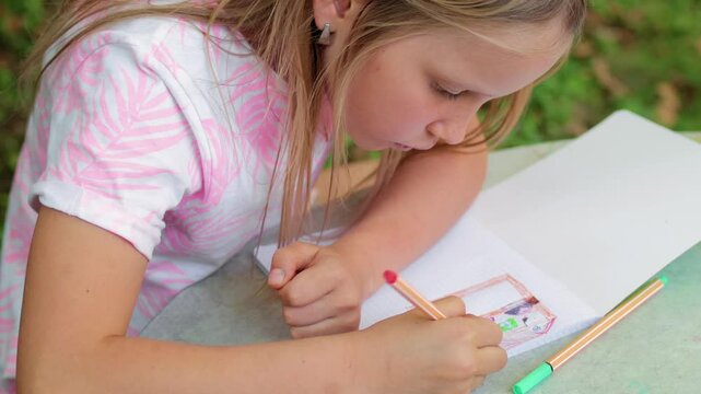 A young girl sits at a table in a summer garden, concentrating while drawing with a fineliner in a graph-paper notebook, expressing creativity through detailed sketch