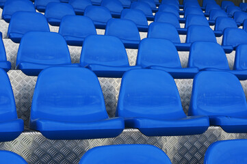 A close-up photograph of empty blue plastic stadium seats arranged in symmetrical rows on a metallic, textured floor © Mikhail Olykainen