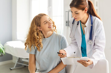 Fototapeta premium Happy smiling young woman patient talking with friendly female doctor therapist holding report file with appointment sitting in office during medical exam in clinic. Medicine and health care concept