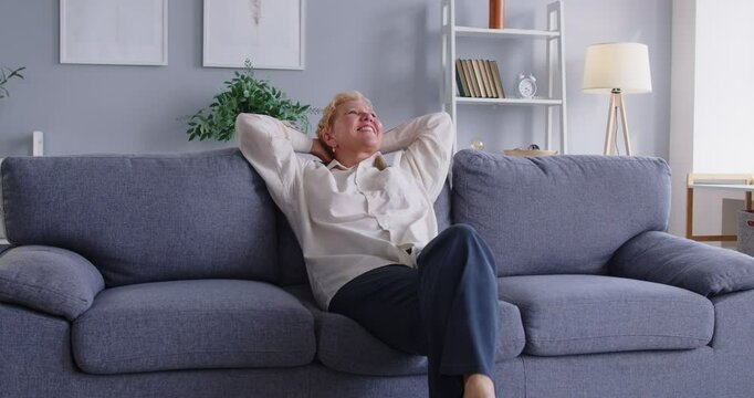 Joyful senior woman, possibly a grandmother, falling back onto a sofa, resting and relaxing at home. She appears happy and content, enjoying a moment of satisfaction in a positive setting.