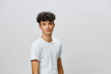 Portrait of a young man with curly hair standing against a plain gray background, wearing a casual white t shirt and showcasing a friendly expression