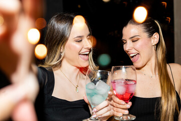 Two women in their 20s and 30s are having fun at a party. The girls take a photo with their mobile phones while holding glasses of alcohol. Party women concept.