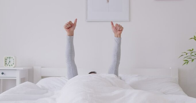 Portrait of faceless male person lying in bed under white blanket stretching his arms and showing thumb up sign. Relaxed sleepy man waking up in the morning with good mood in bedroom. 4k video.