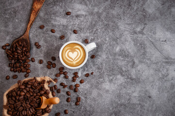 Top view of a hot latte and heart-shaped coffee art in a white cup, a sack, and a wooden spoon with roasted coffee beans, all on a slate table. Space for text.