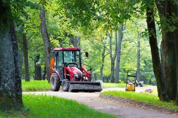 A red tractor with a trailer is parked on a gravel path in a park. Its bucket is filled with gravel. Repairing paths in a park. Preparing for the tourist season.