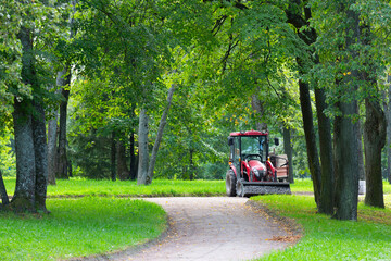 A red tractor with a trailer is parked on a gravel path in a park. Its bucket is filled with gravel. Repairing paths in a park. Preparing for the tourist season.