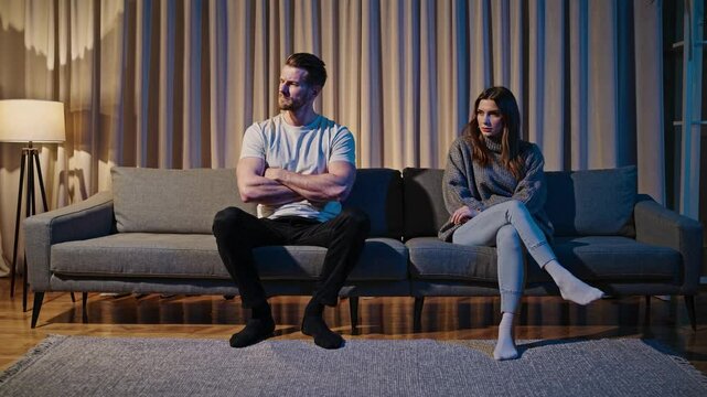 In a warmly lit living room, a couple sits apart on a couch, both looking tense after a disagreement. The atmosphere is heavy as they avoid eye contact, surrounded by soft light