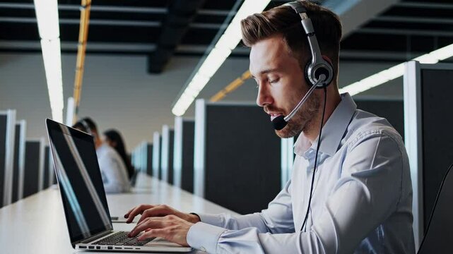 A focused customer service representative wearing a headset types responses on a laptop in a bustling call center. Multiple colleagues work at desks in the background